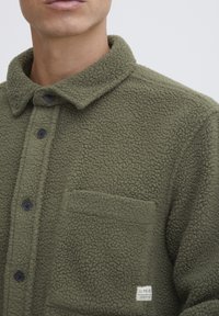 Oliven grønn fleece skjortejakke med kneppet krage, teksturert stoff, og en brystlomme med en etikett. Svarte knapper foran.