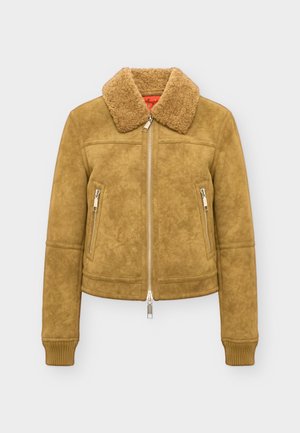 Personne portant une veste en shearling beige avec des poches zippées et un pantalon noir à jambes larges, debout devant un fond clair uni.