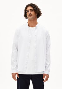 Weißes Langarmhemd mit rundem Kragen, Knopfverschluss und lockerem Schnitt, über einem schlichten weißen T-Shirt getragen.