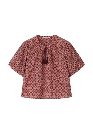 Vrouw draagt een rood topje met patroon, groene short en sjaal, houdt een roze en zwarte gehaakte tas vast, staande voor een beige achtergrond.