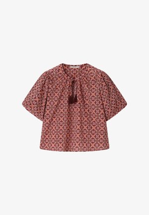 Vrouw draagt een rood topje met patroon, groene short en sjaal, houdt een roze en zwarte gehaakte tas vast, staande voor een beige achtergrond.