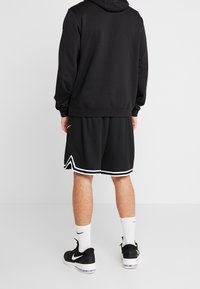 Schwarzer Kapuzenpullover mit Kängurutasche, getragen über schwarzen Sportshorts. Enthält weiße Akzente und Socken, kombiniert mit schwarzen Sneakers.