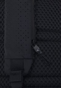 Rucsac negru din material texturat, cu un design perforat, dotat cu un fermoar neted și o curea din țesătură. Accentele notabile includ un mici tragere pentru fermoar.