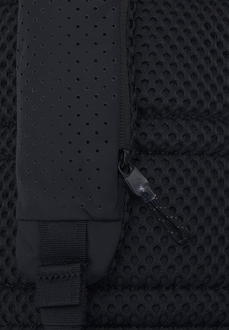 Sac à dos noir en matériau texturé avec un design perforé, doté d'une fermeture éclair lisse et d'une bandoulière en tissu. Les éléments distinctifs comprennent un petit tirette de fermeture éclair.