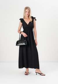 Vestido maxi preto com decote em V, corpete ajustado, cintura franzida e alças com laço. A modelo segura uma pequena mala preta e usa sandálias pretas com tiras.