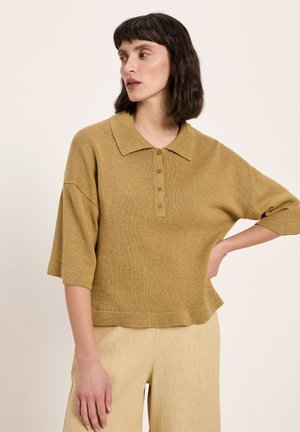 Frau mit dunklen, schulterlangen Haaren, die ein senffarbenes, gestricktes Poloshirt und beige Hosen trägt, schaut nach links und hat die Hand in die Hüfte gestützt.