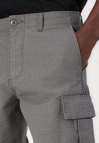 Pantaloni cargo gri cu o textură netedă, având buzunare laterale și un sistem de închidere cu nasture. Cusături vizibile pe margini.
