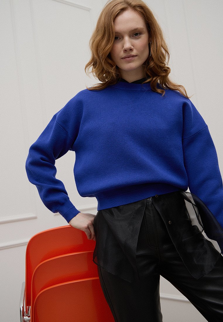 Niebieska bawełniana bluza z wysokim dekoltem i opadającymi ramionami, zestawiona z czarnymi satynowymi spodniami z falbaną przy dołach.