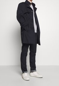 Marinblå trenchcoat i vattentåligt material, figursydd design med knappar, sida fickor, bärs över en ljusgrå sweatshirt, matchad med mörka jeans och vita sneakers.