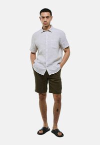 Chemise en lin gris clair à manches courtes avec une poche poitrine, associée à un short vert olive et des sandales glissantes noires, posée contre un fond blanc.