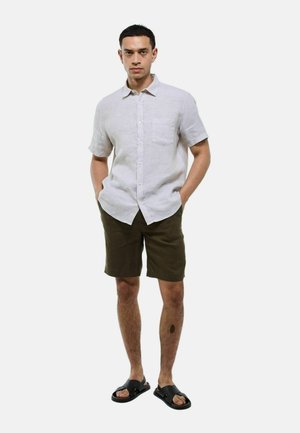 Chemise en lin gris clair à manches courtes avec une poche poitrine, associée à un short vert olive et des sandales glissantes noires, posée contre un fond blanc.