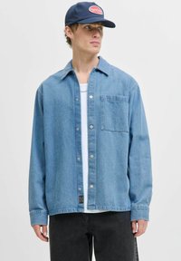 Lichtblauwe denim shirt met een knoopsluiting aan de voorkant, lange mouwen en een borstzak. G gedragen over een wit T-shirt, gecombineerd met donkere jeans.