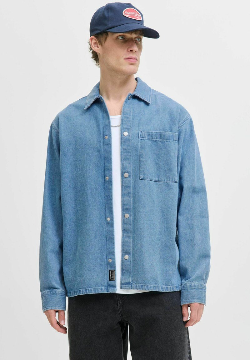Lichtblauwe denim shirt met een knoopsluiting aan de voorkant, lange mouwen en een borstzak. G gedragen over een wit T-shirt, gecombineerd met donkere jeans.