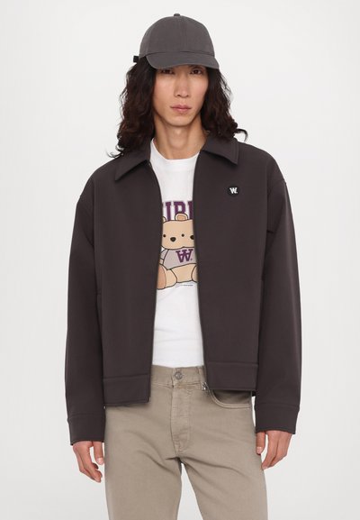 Pruun sweatshirti jope, millel on terav krae, lukuga ja logomärk, kantud valge graafikaga T-särgi ja beeži pükstega. Hall müts.