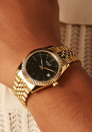 Goldene Armbanduhr mit schwarzem Zifferblatt, geriffelter Lünette und strukturiertem goldfarbenem Armband. Verfügt über Stundenmarkierungen und eine Datumsanzeige bei 3 Uhr.