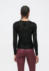 Schwarzes Langarmshirt mit strukturiertem Blumenmuster aus transparentem Stoff, kombiniert mit glänzenden, eng anliegenden bordeauxroten Hosen.