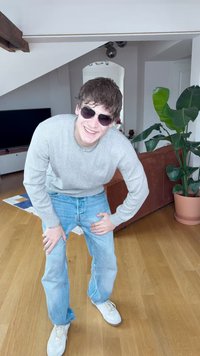 Camisola cinza clara, jeans azuis e sapatilhas brancas, acompanhados por óculos escuros. Em pé sobre chão de madeira com plantas verdes ao fundo.