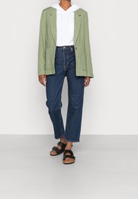 Blazer verde in lino, felpa bianca con cappuccio, jeans a vita alta blu e sandali neri. L'outfit presenta una vestibilità rilassata e un design minimalista.