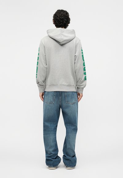 Person with curly hair stands facing away wearing a gray hoodie with green text on sleeves and loose blue jeans against a white background.