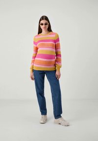 Woman standing wearing striped pink, orange, and yellow sweater, blue jeans, beige sneakers, and oval sunglasses against plain background.