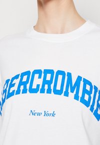 Weißes Baumwoll-T-Shirt mit fettem blauen Text "ABERCROMBIE" im Bogen-Design und kleinerem "New York" darunter. Runder Halsausschnitt, kurze Ärmel.