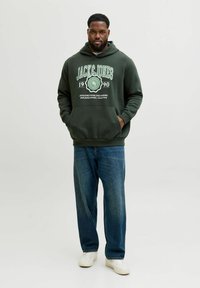 Sudadera verde oscura con bolsillo frontal y texto gráfico blanco, combinada con jeans sueltos azules y zapatillas blancas. Diseño simple, estilo casual.