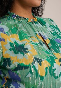 Blommig blus i grönt, gult och blått med rynkad halsringning och ett nyckelhål fram. Har glänsande vertikala ränder över tyget.