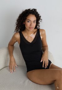Black tank top with lace trim, fitted design, and soft fabric. Model seated on a light, textured surface against a neutral background.