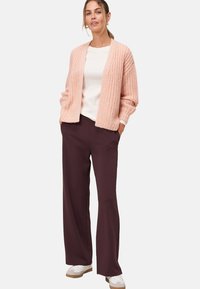 Cardigan rosa lavorato a maglia con vestibilità ampia, texture a coste, indossato sopra un top crema e abbinato a pantaloni a gamba larga marrone scuro e scarpe bianche.