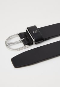 Juodas odinis diržas su sidabriniu metaliniu sagtimi, išgraviruota "Calvin Klein". Diržo juostelė turi sklandų paviršių ir švarų dizainą.