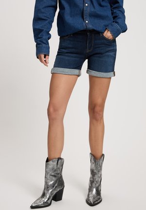 Dark blue denim shorts with rolled cuffs, paired with metallic silver cowboy boots featuring a stacked heel and pointed toe.
