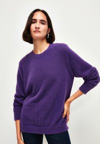 Lila, flauschiger Pullover mit rundem Ausschnitt und langen Ärmeln. Gerippter Saum und Bündchen, getragen über dunklen Jeans, vor einem schlichten Hintergrund.