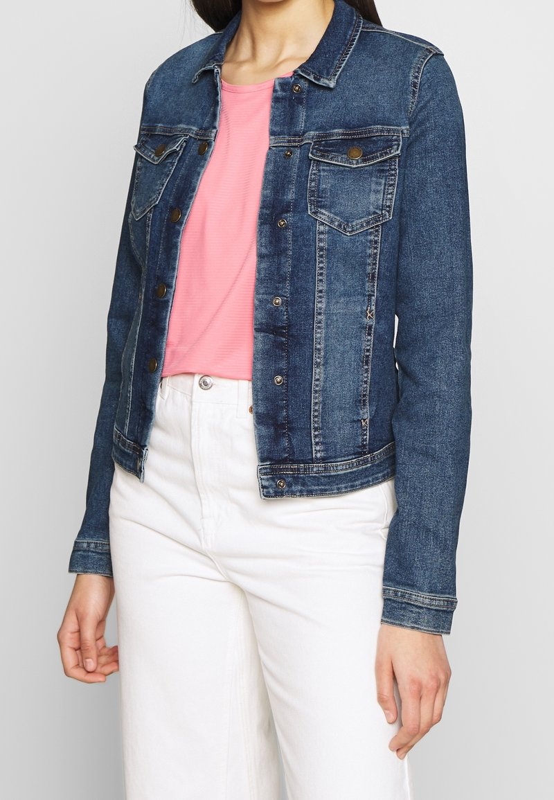 Tammikatu tummansinisessä denim-takissa, jossa on rintataskut, etupainikkeet ja lyhyt leikkaus, päällä vaaleanpunainen paita ja valkoiset housut.