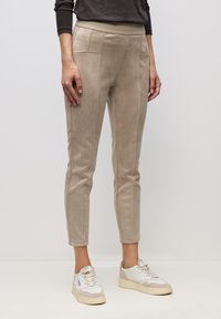 Osoba oblečená v béžových úzkých kalhotách s viditelnými švy a kapsami, v kombinaci s bílými a béžovými teniskami, stojící před jednoduchým bílým pozadím.