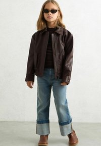 Pruun kunstnahast jope, millel on veidi läikiv tekstuur, koos kerge sinise teksapükste ja pruuni pahkluu saapaga. Mustad prillid täiustavad välimust.