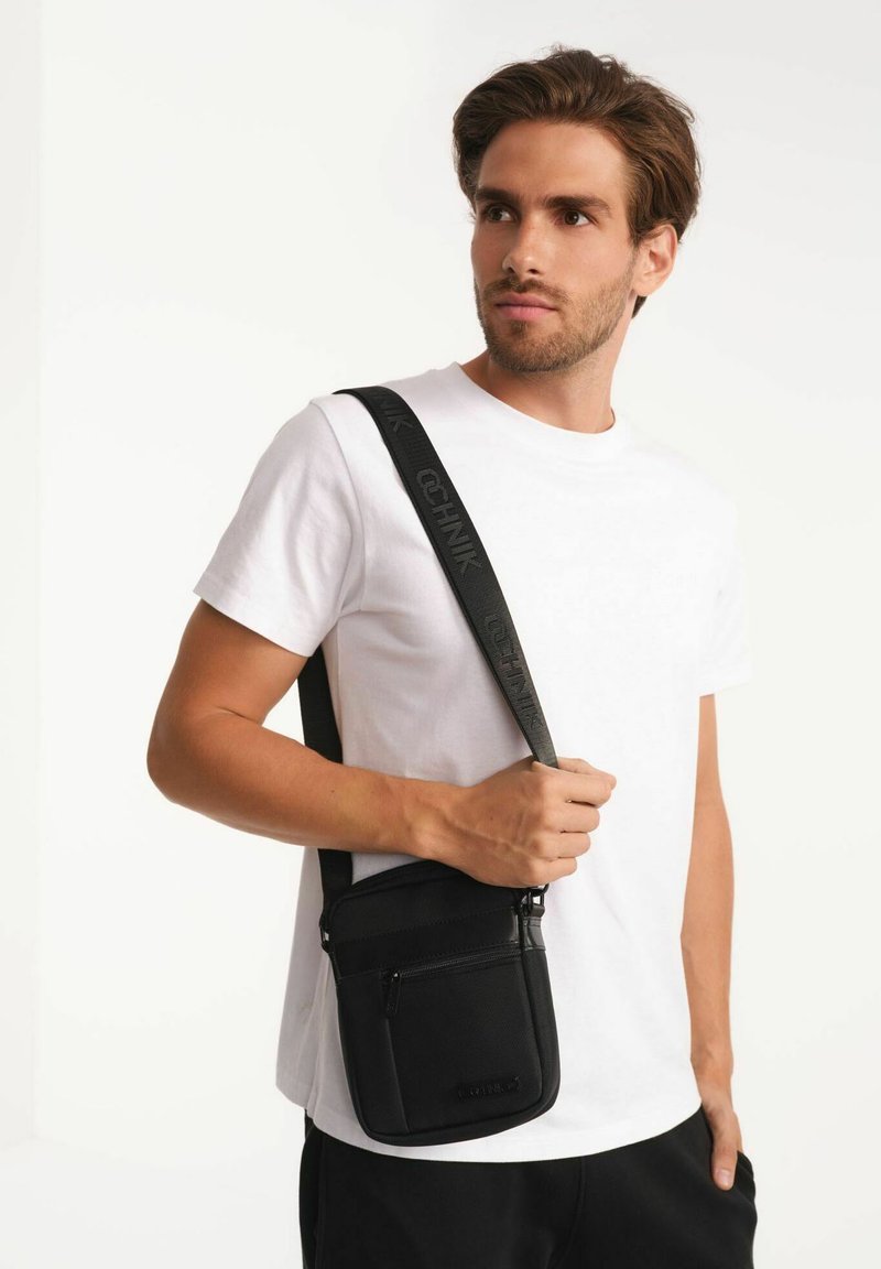 Czarna torba crossbody o teksturowanej powierzchni, regulowany pasek z logo oraz zasuwane przegródki dla bezpiecznego przechowywania.