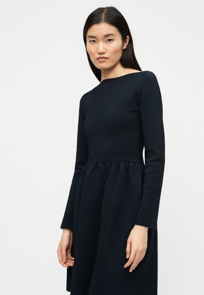 Φόρεμα με μακριά μανίκια σε σκούρο μπλε χρώμα με υφή ριγέ. Το εφαρμοστό μπούστο καταλήγει σε μια μέση με πιέτες που δημιουργούν φούστα με φουσκωτή γραμμή.