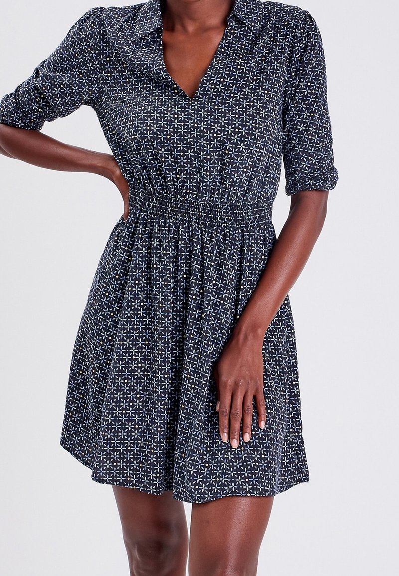 Femme portant une robe à motifs bleu foncé avec un décolleté en V, une taille froncée et des manches retroussées, se tenant devant un fond clair uni.
