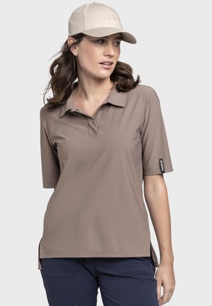 Frau mit beiger Mütze und taupefarbenem kurzärmeligem Poloshirt, die seitlich schaut, mit marineblauer Hose vor schlichtem Hintergrund.