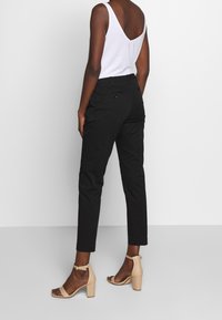 Kvinna iklädd svarta slim-fit ankelnbyxor, vit ärmlös topp med V-ryg och beige klacksandaler, stående mot en enfärgad vit bakgrund.