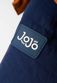 Prim-plan al unei secțiuni de țesătură bleu marin cu o petrecere rectangulară cusută, care are inscripția "JoJo maman bébé" în litere albe.