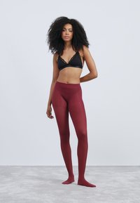 Bordeauxfarbene Strumpfhosen mit glatter, transparenter Textur, getragen mit einem schwarzen Bralette mit Spitzenbesatz. Das Model steht auf einer grauen Fläche vor einem hellen Hintergrund.