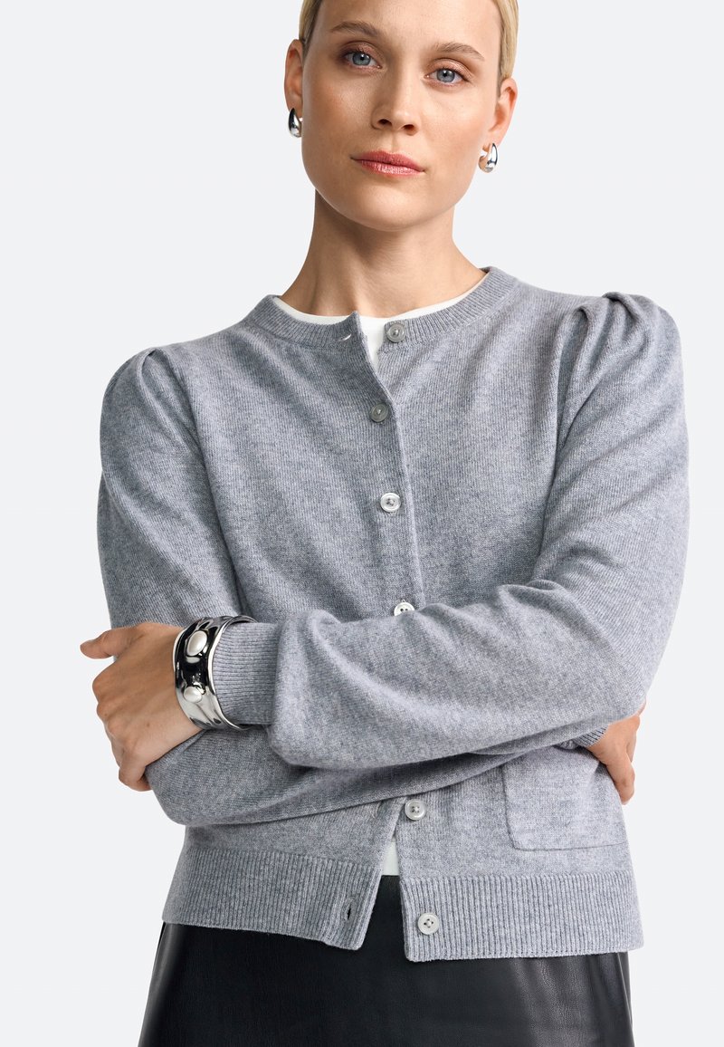 Femme portant un cardigan gris à boutons et un bracelet en argent, les bras croisés, se tenant contre un fond clair.