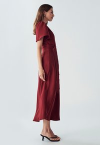 Robe midi en bordeaux profond, avec des manches bouffantes, une taille cintrée avec un détail en nœud, et une jupe fluide. Le matériau a une texture lisse.