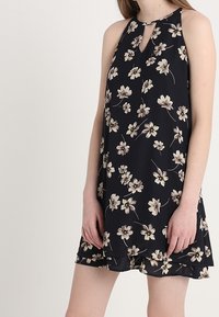 Kwiatowa sukienka bez rękawów z czarnego materiału, z dekoltem typu keyhole i beżowymi wzorami kwiatowymi na całej powierzchni; luźny krój z delikatnym rozszerzeniem.