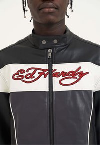 Giacca in pelle bianca e nera con colletto alto, caratterizzata da testo ricamato rosso "Ed Hardy" sulla parte anteriore, hardware argentato e chiusura a zip.