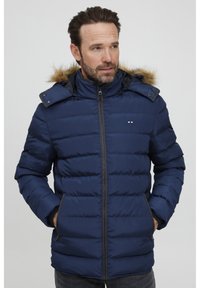 Mørkeblå puffjakke med hette foret med falsk pels, vertikal quilting, to frontlommer og glidelås foran.