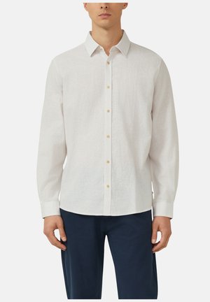 Homme portant une chemise blanche à manches longues boutonnée avec col et un pantalon bleu marine, debout devant un fond uni.