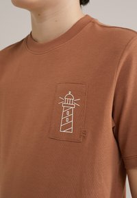 Ruda medvilninė marškinėliai su balta švyturio grafika ant kišenės. Jie turi raištinį kaklą ir mažą logotipą apatiniame dešiniajame kampe.