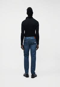 Άνδρας με κοτσίδες ντρέντλοκ, φοράει μαύρο μακρυμάνικο μπλουζάκι, μπλε τζιν και μαύρα παπούτσια, στέκεται με την πλάτη προς το φακό σε απλό λευκό φόντο.